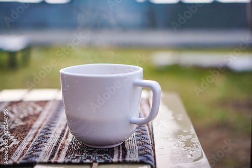 Cuadro en lienzo White ceramic mug sits on a detailed patterned cloth outdoors releasing wisps of hot steam into the cool air while raindrops settle on the nearby surface during a peaceful morning