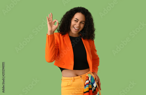 Stylish young African-American woman in orange blazer showing OK on green bac...