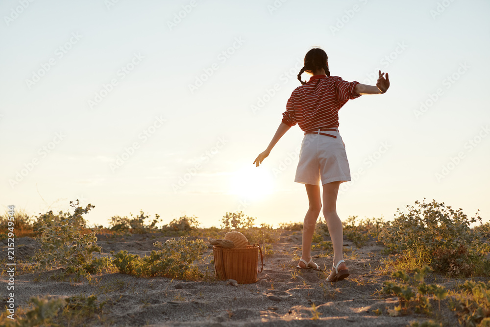 Obraz premium Young woman with braided hair walking barefoot on sandy terrain at sunset during a summer picnic. The female enjoys a peaceful lifestyle while exploring the golden light in a warm outdoor setting.