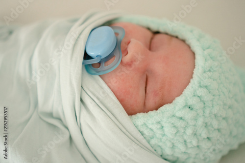 A newborn baby sleeps peacefully indoors, sucking a pacifier with a strong reflex while swaddled in white. The calm infant rests soothed in closeup, safe and comfortable in soft nursery light.