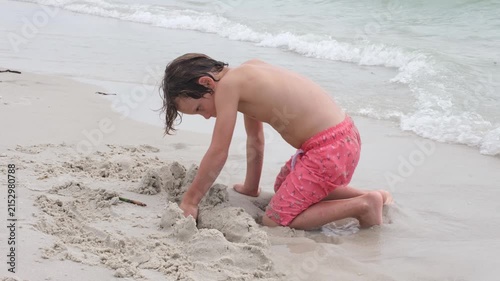 A young boy kneels on the shoreline while digging and shaping wet sand beside gentle ocean waves. The candid beach scene highlights childhood creativity, outdoor recreation, and imaginative play.