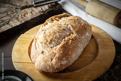 Konstfotografi Freshly baked artisan sourdough bread loaf resting on a wooden board in a rustic kitchen setup with flour, a rolling pin, and traditional baking equipment