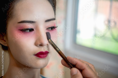 Close-up unrecognized makeup artist applying traditional Japanese geisha cosmetics on female model with white face foundation, red eyeshadow and vivid red lipstick