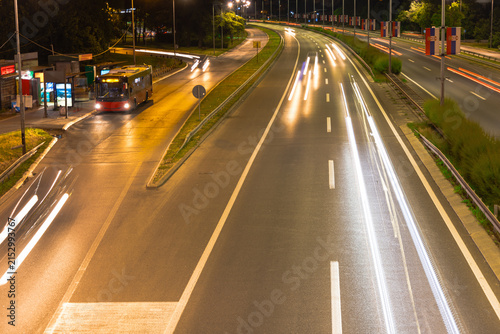 High-angle night traffic scene on a multi-lane highway with long exposure light trails from vehicles, a parked bus at a station, and glowing street lamps illuminating the wet asphalt road.