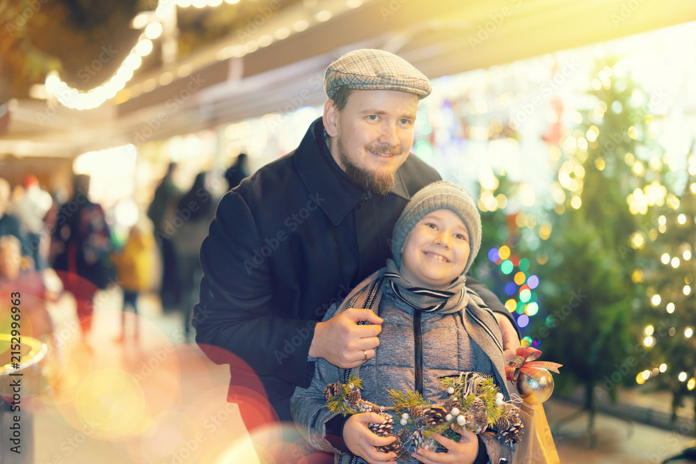 Obraz premium Boy with father stand in middle outdoor New Years shopping malls, dreamily imagines Christmas holiday. They stared at beautiful goods in window of fair, enthusiastically watching something fascinating
