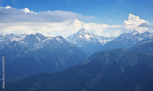 Landscape of high rocky mountains focusing on a majestic central snowy peak above dark green ridges under a blue sky with fluffy white clouds.