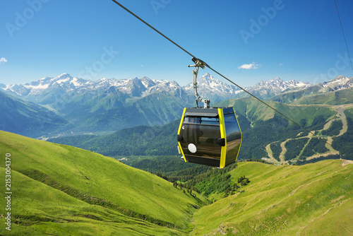 Modern yellow cable car cabin moving on a ropeway above lush green alpine hills towards high rocky mountains with snowy peaks under a clear blue sky. Travel infrastructure concept design element 