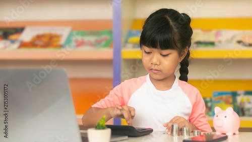 Cute Asian girl using calculator with stack of coins, laptop and piggy bank for financial education concept