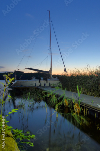 Fotografía Sailboat docked at sunset with calm water and reflecting sky in a tranquil setti