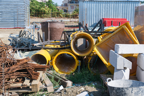 A haphazard pile of bright yellow industrial cylindrical tubes and construction equipment rests on the grass alongside metal scaffolding and rusty rebar at an outdoor building site.