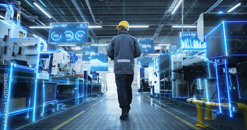 Foto Rear View of an African American Male Worker Walking Between Machines in a Digitalized Factory as Glowing Holographic Panels Display Line Efficiency Percentages, Bar Graphs and Ai Diagnostic Scans