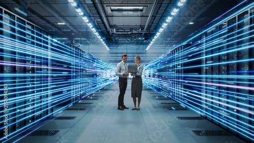 Two Caucasian It Professionals, a Man and a Blonde Woman, Review Network Data on a Laptop While Standing Between Server Racks Emitting Fast Neon Blue Fiber Optic Light Trails.