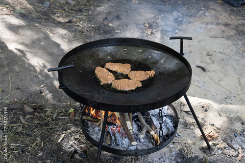 Delicious meat cutlets frying in a large round cast iron pan over an open campfire with wood and ash outdoors, perfect for camping, picnic, and rustic cooking adventures in nature.