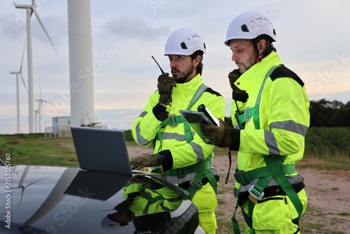 Team engineers are reviewing data on laptops and discussing maintenance at a renewable energy wind farm. The background is a field of wind turbines. One person was using a radio to communicate.