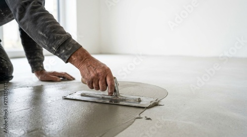 Worker spreading self-leveling cement compound on floor with trowel.