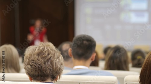 Audience listens to speaker in conference room. People sit attentively, facing presentation screen. Focus on back of heads, suggesting engagement. Ideal for business, education. Suitable for training
