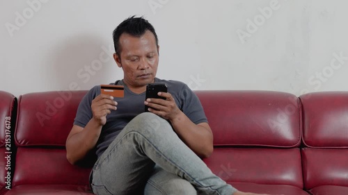 Asian man at home sitting in sofa. Male client banking buying shopping online with credit bank card and mobile smartphone, e-commerce order delivery payment internet pay.