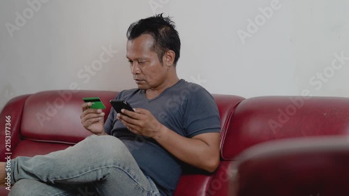 Asian man at home sitting in sofa. Male client banking buying shopping online with credit bank card and mobile smartphone, e-commerce order delivery payment internet pay.