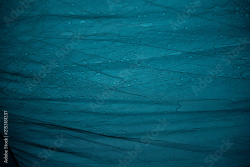 A wide close-up of water drops resting on a vibrant blue, wrinkled waterproof tent tarp. The image highlights the texture of synthetic material, folds, and hydrophobic surface during a rainy day outdo