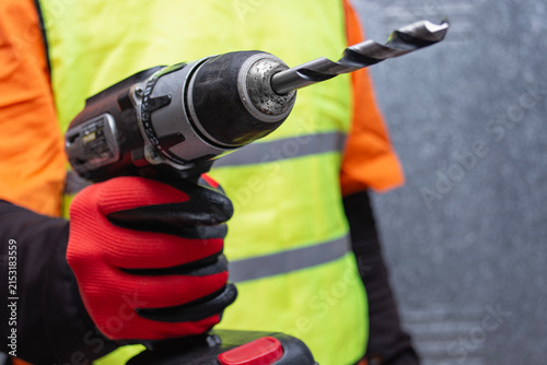 Foto Construction worker in reflective high-visibility vest and protective gloves holding cordless electric power drill with large drill bit