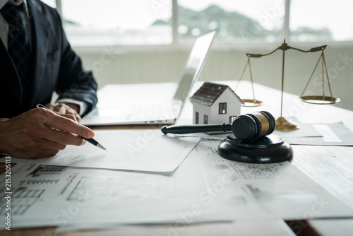 Real estate professional reviewing property documents at a desk with a house model, legal gavel, architectural plans, laptop, and scales, representing property law and transactions.