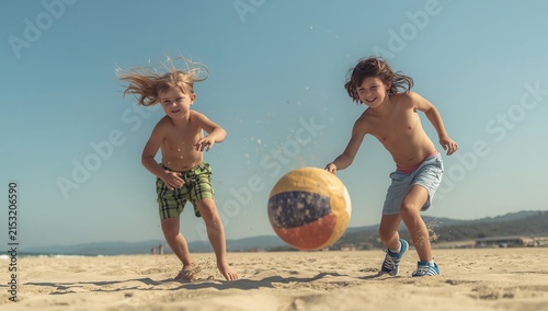 Two boys playing with a ball on a sandy beach under a clear blue sky