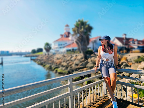 A stylish female tourist wearing a cap relaxes on a wooden boardwalk railing, enjoying the beautiful view of  Long Beach bay in California with a blue ocean and sky. 