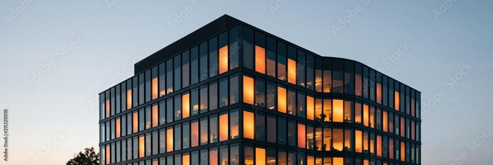 Fototapeta premium Contemporary multi story glass office building at dusk with warm interior lighting for architectural wallpaper and design background
