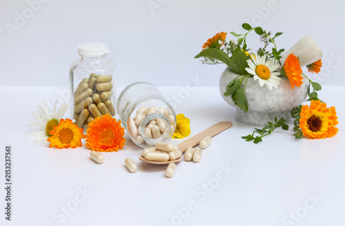 Two glass bottles and wooden spoon with capsules of herbal food supplements on white background. Nearby stone mortar with fresh medicinal herbs, calendula, chamomile, thyme leaves and Kuril tea