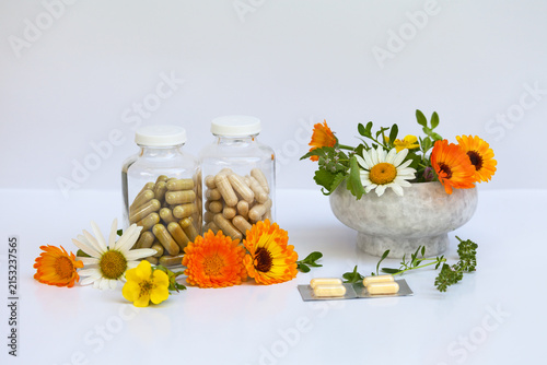 Two glass bottles with capsules of herbal dietary supplements on white background. Nearby is mortar with medicinal herbs: calendula, Kuril tea, chamomile, thyme leaves. Alternative medicine
