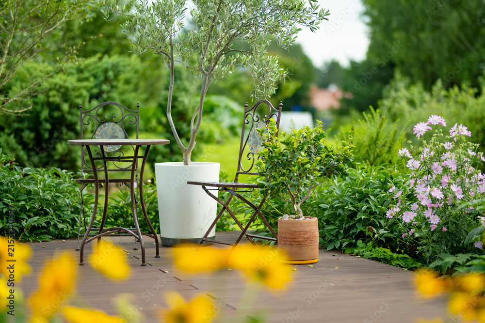 Fototapeta premium Small table and chairs stand on a wooden garden terrace surrounded by lush greenery and potted plants. Cozy outdoor seating area creates a peaceful summer retreat.