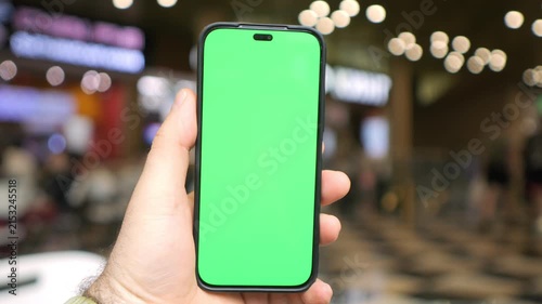 Hand holds a smartphone with green screen. Phone display is blank and ready. Background features a blurred mall. People move through the space. Footage ideal for mobile content