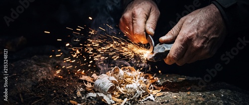 Outdoorsman igniting fire with flint and steel creating golden sparks hitting dry birch bark tinder for primitive wilderness survival camp