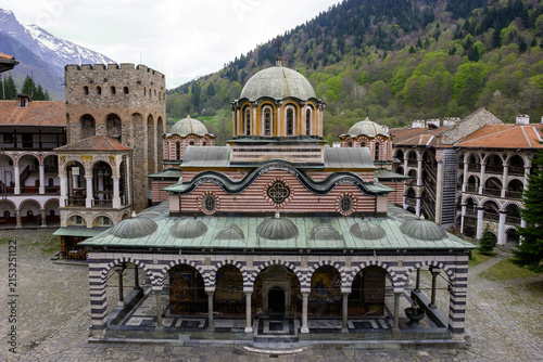 фотографія The Rila Monastery, a UNESCO World Heritage site in Bulgaria, is seen with its iconic church and surrounding monastic buildings, set against a backdrop of snow-capped mountains and lush green forests