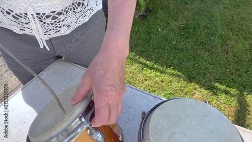 Two handcrafted hand drums with natural animal-hide drumheads rest on a table outdoors. A woman carefully cleans the smaller drum with compressed air, Close-up static shot
