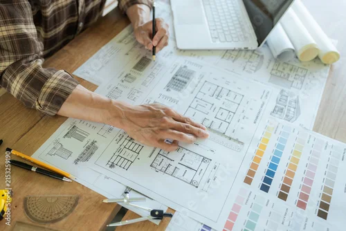 Fototapeta na wymiar Architect drawing and reviewing detailed building blueprints on a desk with a laptop, rolled plans and color samples, representing architectural design and construction planning.