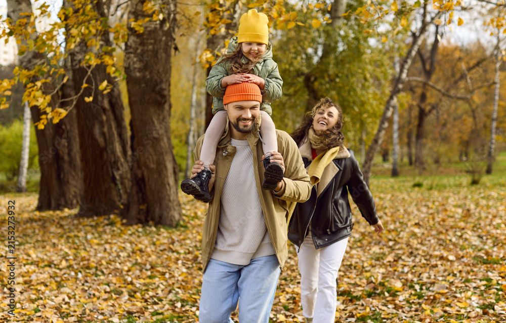 Obraz premium Autumn couple play with leaves in park. Happy parents carry their child on shoulders, enjoying nature and golden foliage during a relaxed stroll. Joyful fall family bonding concept in nature.
