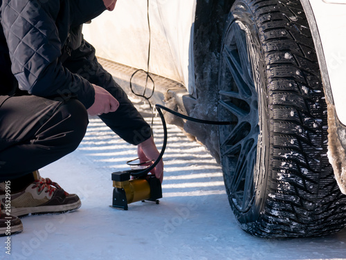 Man inflating a snow-covered car tire with a portable air compressor in winter. Roadside vehicle maintenance, travel safety and emergency assistance concept.