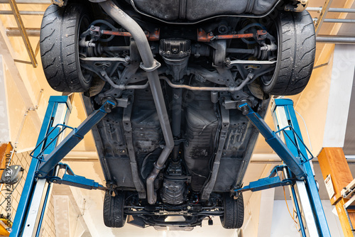 Wide view from directly below showing the complete chassis, exhaust system, and two-post hydraulic lift supporting a race car.
