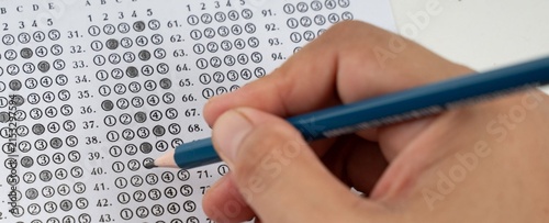 Hand marks answer bubbles on a standardized test sheet. Blue pencil glides over rows of multiple-choice options. Fingers grip the tool with purpose and focus