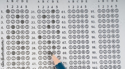 Hand points to numbered grid of black and white circles. Grid shows rows labeled with letters and numbers. Each circle is filled or empty, forming a pattern. Fingers gesture toward specific entries
