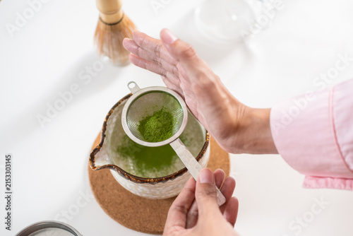 Obraz na plátně Top view of hands tapping a stainless strainer to sift fine organic matcha powder into a ceramic bowl