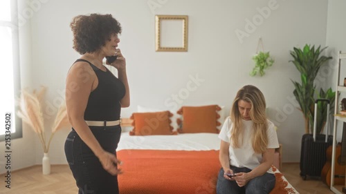 Woman in black sleeveless top holds smartphone to ear while another woman sits on bed with orange throw in building; tension estrangement.