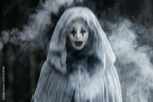 Spooky and scary Halloween themed image in a dark forest with smoke and an eerie woman with a mask. .