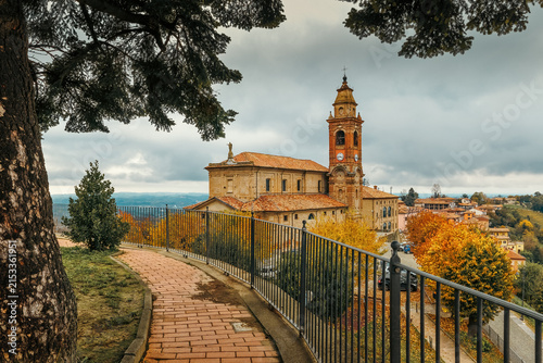 A church under an overcast sky on a small town square surrounded by autumnal trees, in Diano d'Alba, Italy.