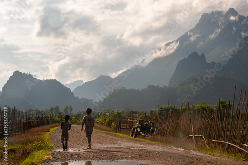 VANG VIENG, LAOS – Two boys walk along a dirt road toward misty karst mountains near Vang Vieng, a scene of everyday rural life amid the dramatic limestone landscape.
