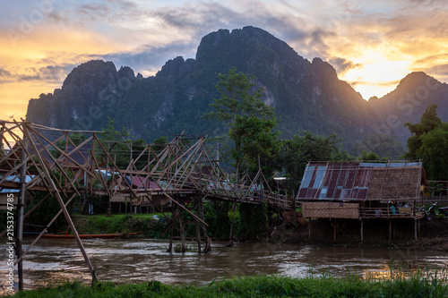 VANG VIENG, LAOS – A bamboo walking bridge spans the river near Vang Vieng at twilight, its silhouette framing the karst mountains against a colorful sunset sky.