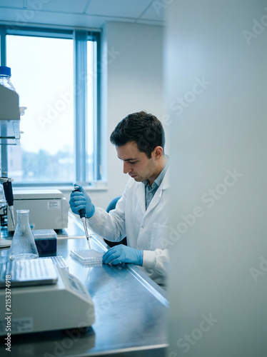 A man wearing a lab coat and blue gloves is using a pipette to dispense liquid into a multiwell