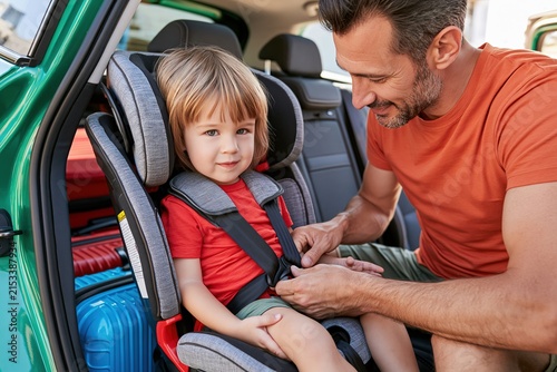 Father fastening a young boy into a child car seat with luggage visible in the vehicle, ideal for road trips, parenting safety and family travel concepts.