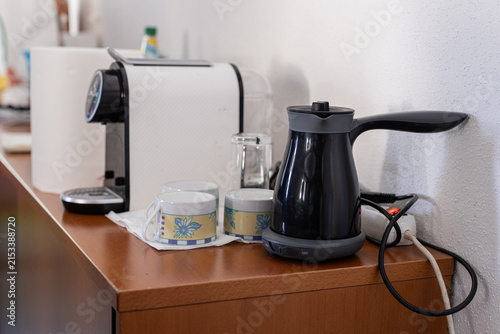 A black electric Turkish coffee maker sits on a wooden countertop alongside small floral espresso cups, glasses, and a white coffee capsule machine in a home kitchen setting.